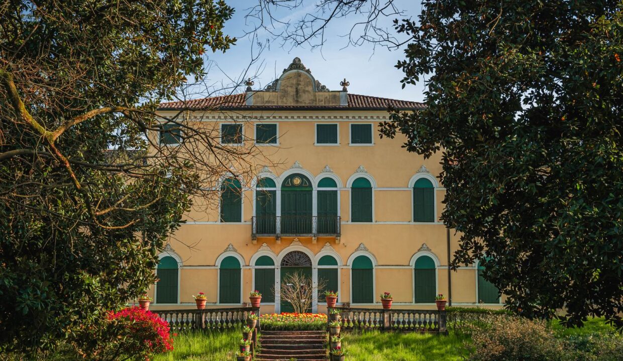 Facciata di una villa d'epoca con persiane verdi e scalinata, incorniciata dagli alberi del giardino
