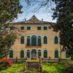 Facciata di una villa d'epoca con persiane verdi e scalinata, incorniciata dagli alberi del giardino