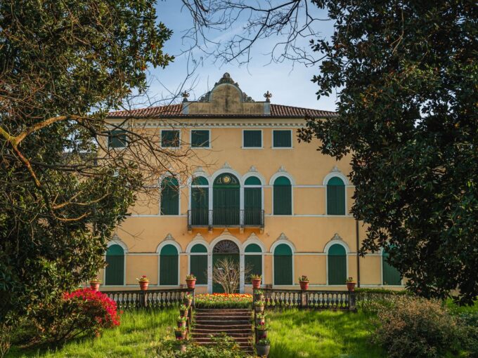 Facciata di una villa d'epoca con persiane verdi e scalinata, incorniciata dagli alberi del giardino